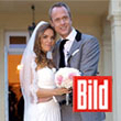 image - Reinweiss Hochzeiten Wedding photo of a couple in front of a building, bride in a white dress with a bouquet of flowers, groom in a gray suit.