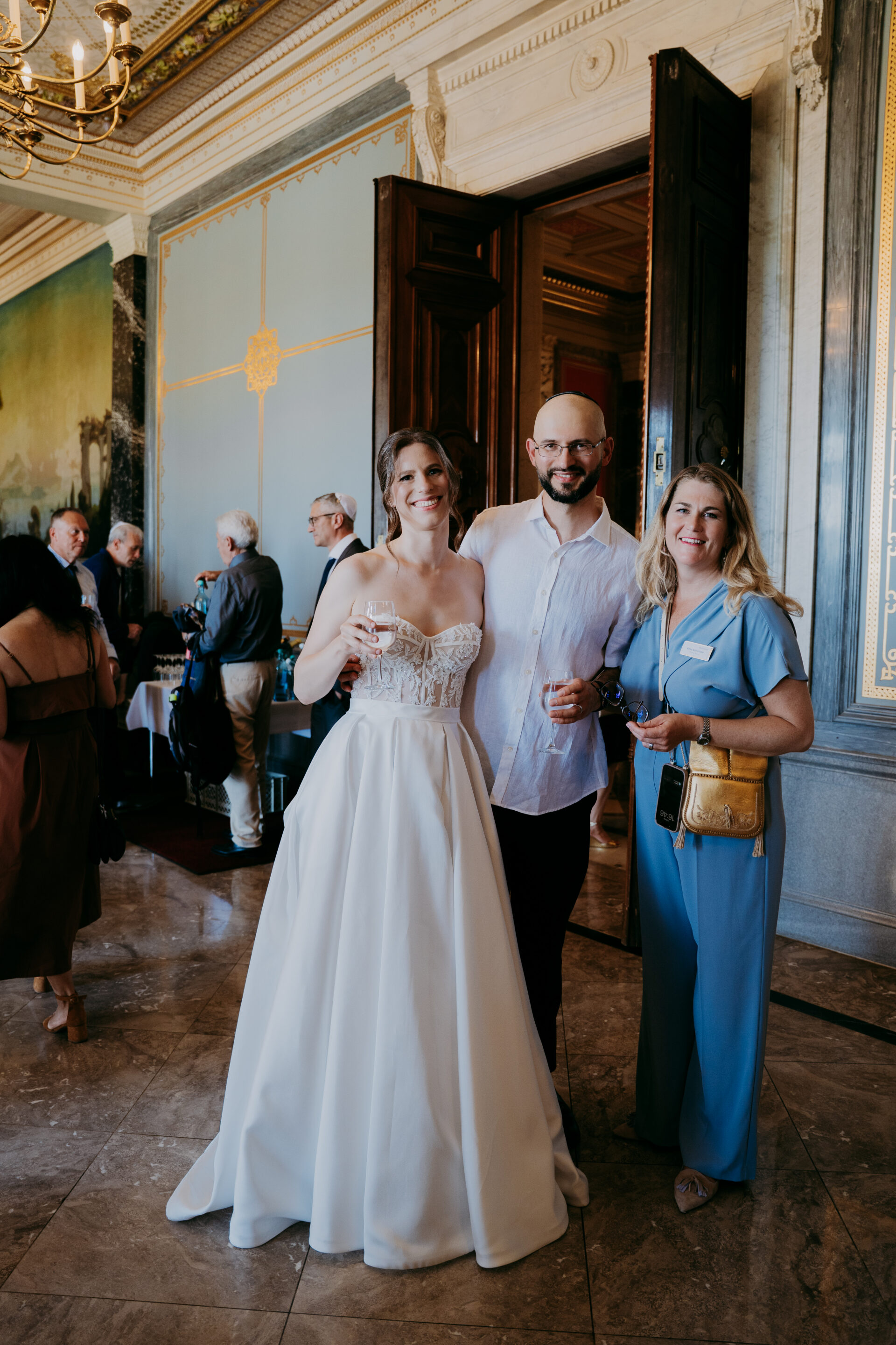 Elena Budinstein_Mariage_Dresde - Reinweiss Hochzeiten Mariée vêtue d'une élégante robe blanche se tenant avec deux invités dans une salle de fête lors d'un mariage à Dresde.