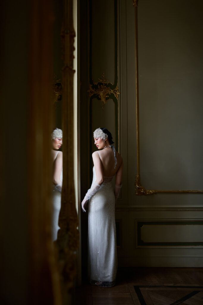 ermelerhaus-micro-wedding - Reinweiss Hochzeiten Элегантная невеста в простом блестящем платье стоит перед зеркалом в роскошной комнате.