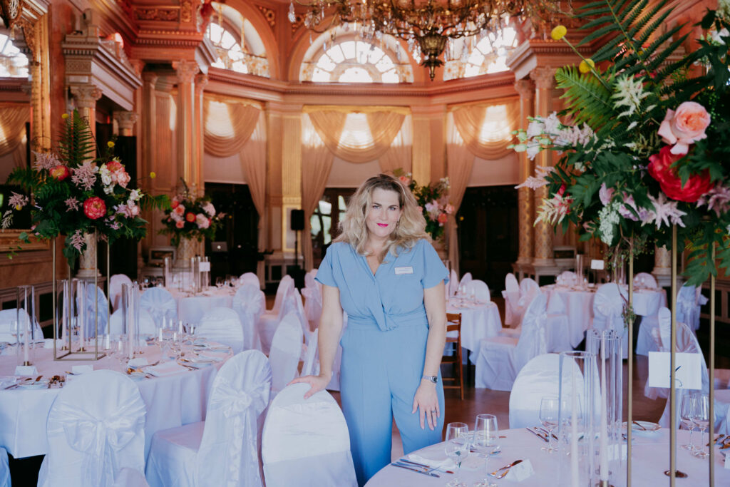 Hochzeitplaner_Berlin - Reinweiss Hochzeiten Elegant dekorierter Hochzeitsraum mit Tischen und Blumenarrangements; eine Frau in blauer Kleidung steht lächelnd im Vord...